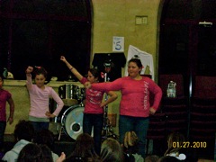 Three girls standing up in front of a crowd with their arms out. There is a drumset behind them. 